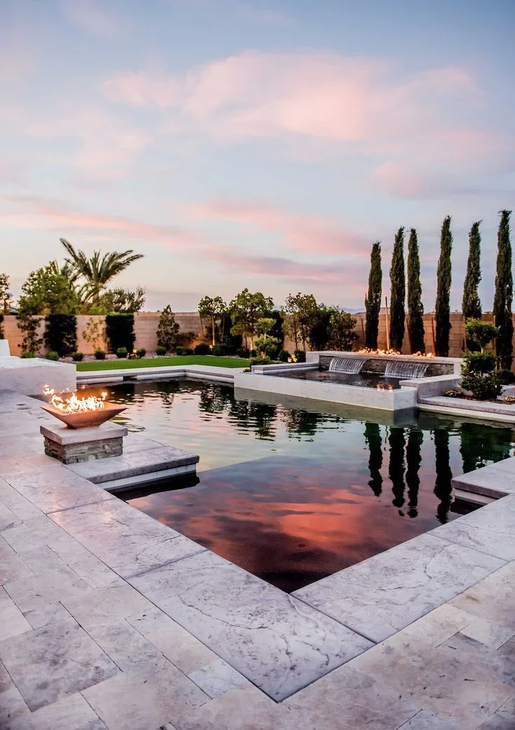 Travertine pool deck with multiple connected pools, water features, and copper fire bowl at sunset