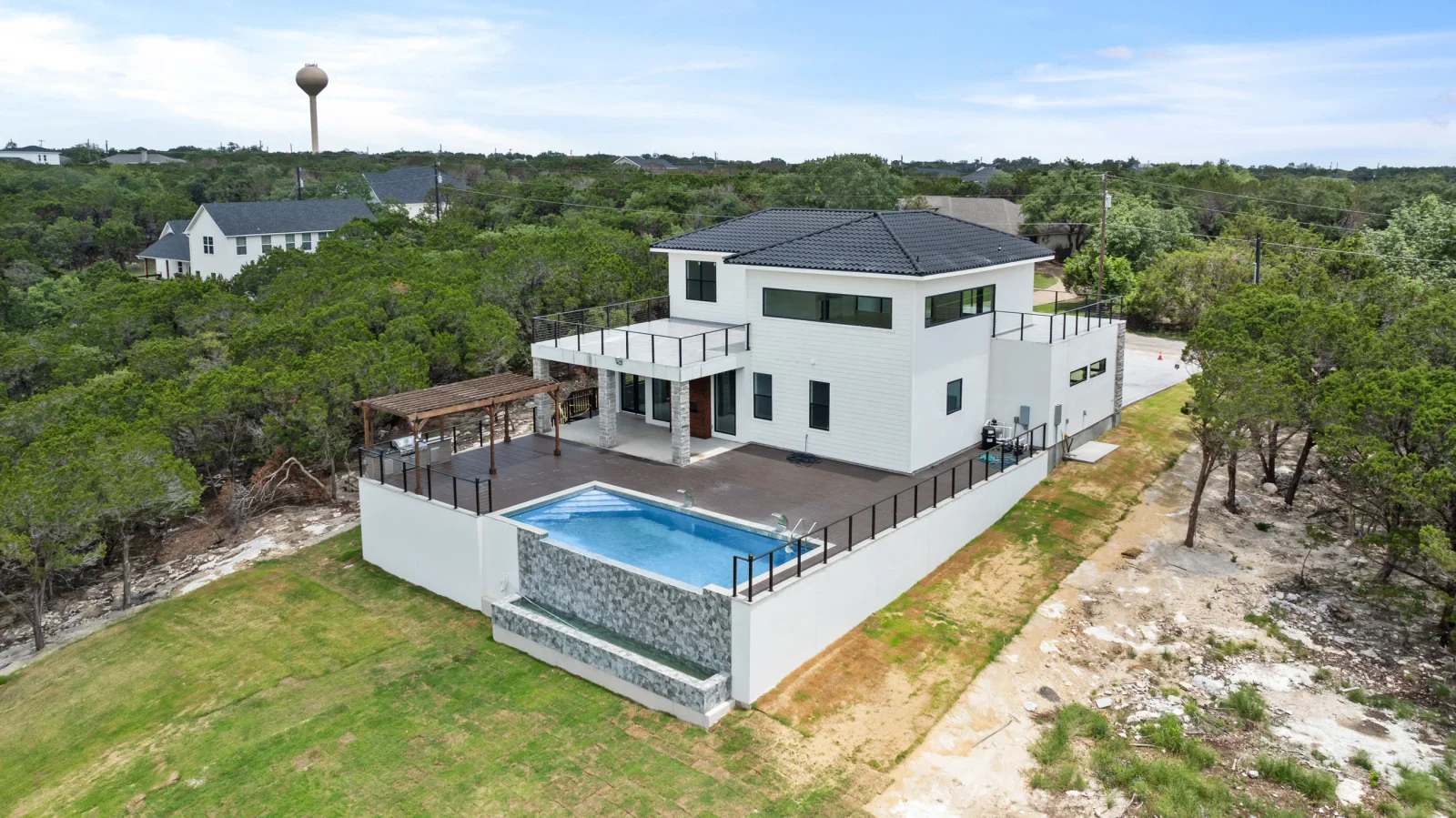 White modern home with dark roof, pool, stone walls, and metal railings on hillside lot