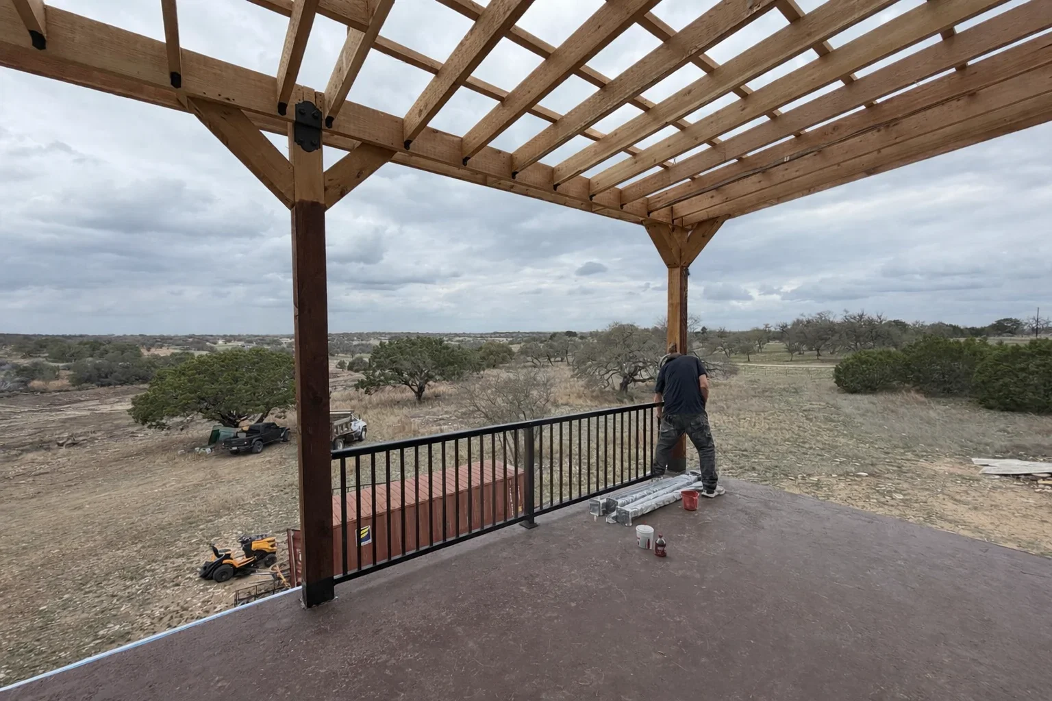 Wooden pergola with lattice roof and black metal railing on concrete pad, rural setting