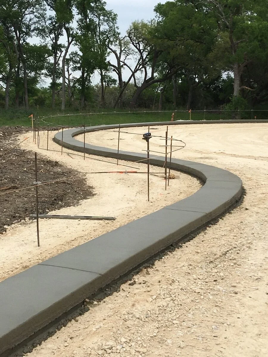 Curved gray concrete curbing on gravel base with reinforcement stakes and protective fencing