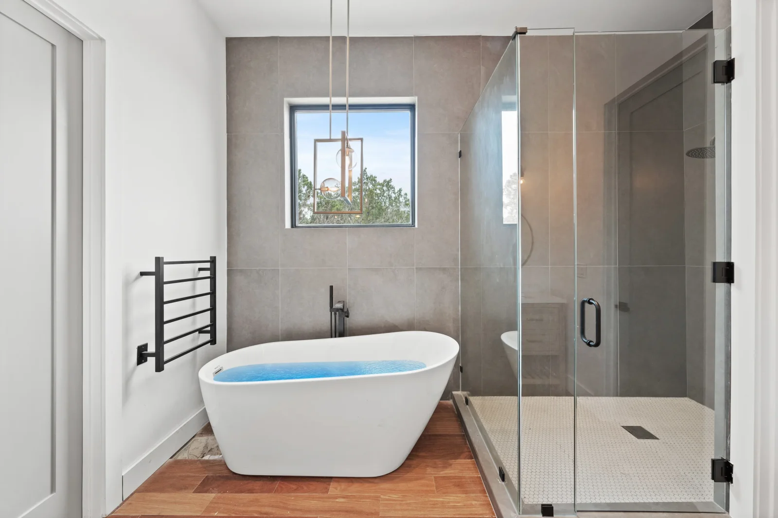 Freestanding white soaking tub, frameless glass shower, large-format tile walls, wood flooring, black fixtures