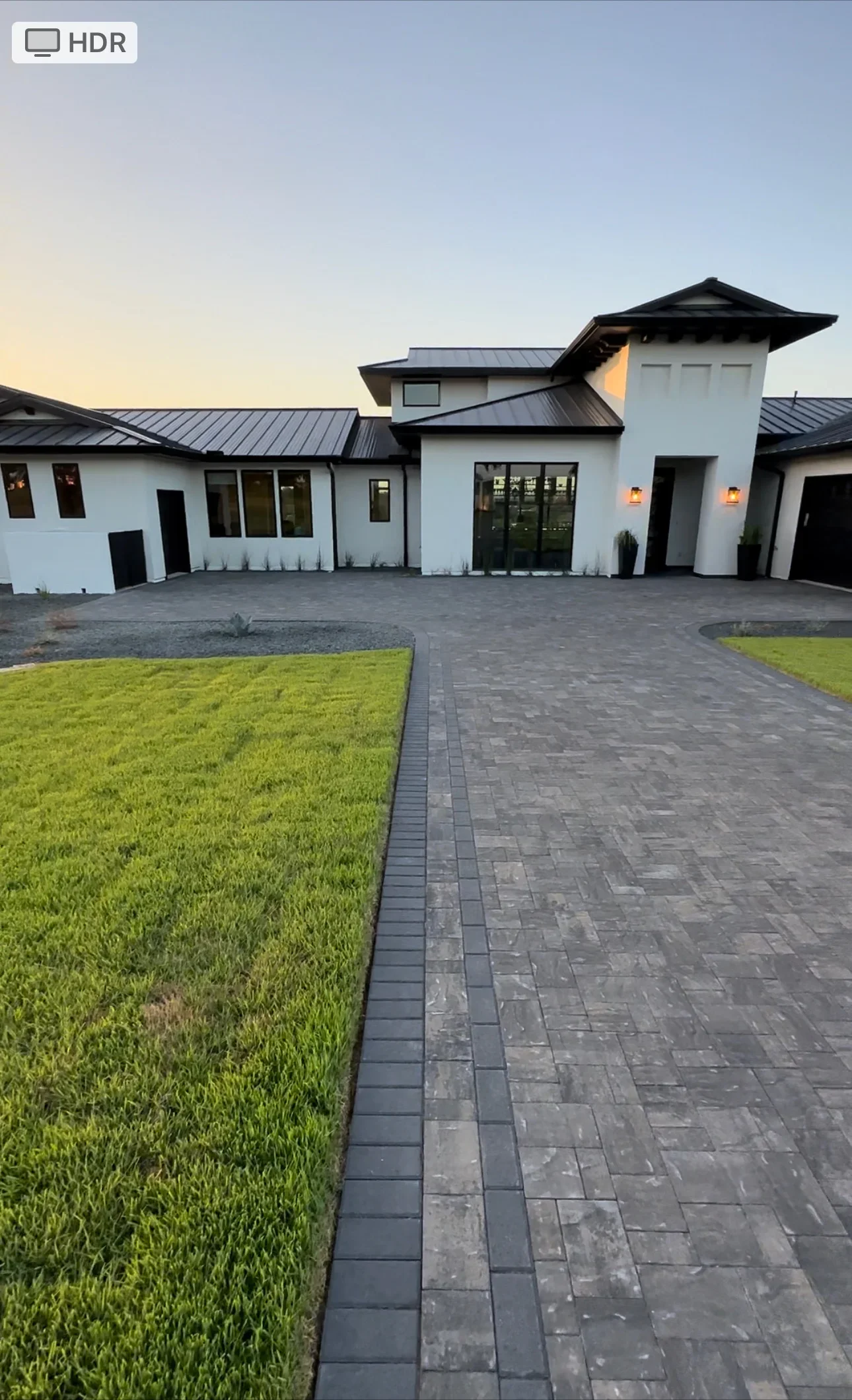 Charcoal paver driveway with black edging, fresh sod lawn, and modern white home with metal roof